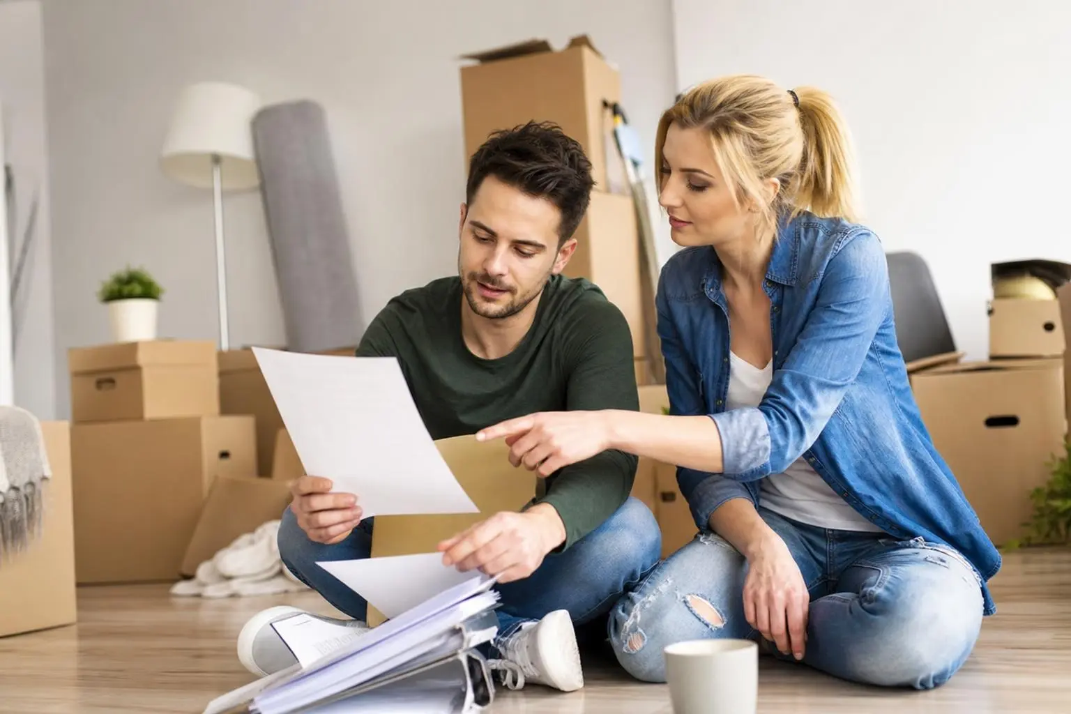 Deux personnes assises par terre dans un logement vide, examinant des documents et des plans autour de cartons, en cours de déménagement. check list déménagement complète leurs démarches.