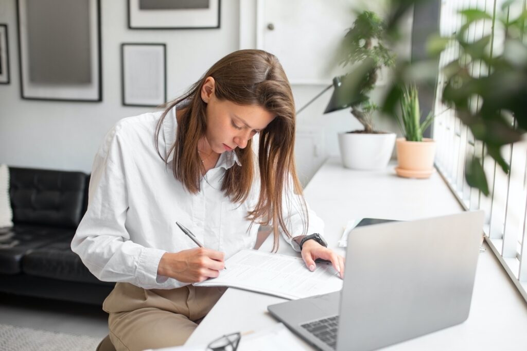 Femme travaillant à domicile: elle remplit des documents sur une feuille près d’un ordinateur portable, en environnement lumineux et épuré. Démarches administratives déménagement.