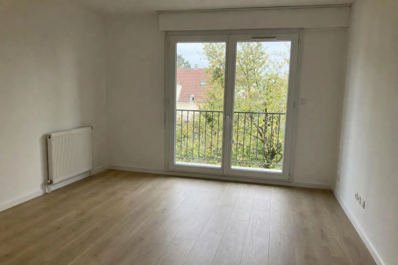 Pièce vide avec parquet clair et grande porte-fenêtre donnant sur un balcon, radiateur à gauche et murs blancs. Inclut une ambiance calme pour le déménagement. Vérifier le checklist déménagement pour organiser les étapes.
